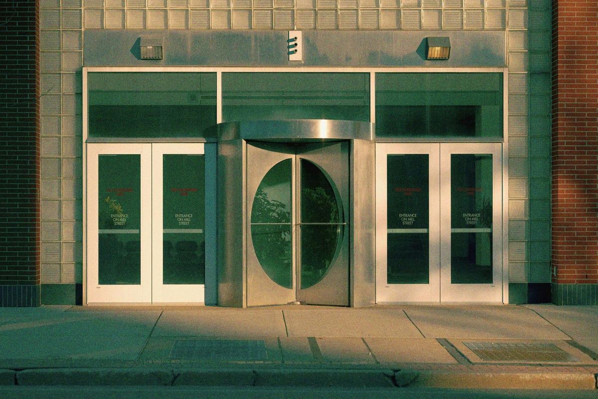 A glass and brick building entrance in warm evening light, with a rounded steel door and no people.
