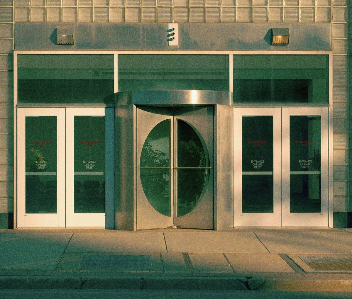 A glass and brick building entrance in warm evening light, with a rounded steel door and no people.